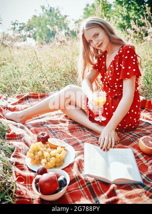 Picnic scene: pretty smiling girl with glass of orange juice sitting on a plaid and reading book in park. Outdoor portrait of beautiful young woman in Stock Photo
