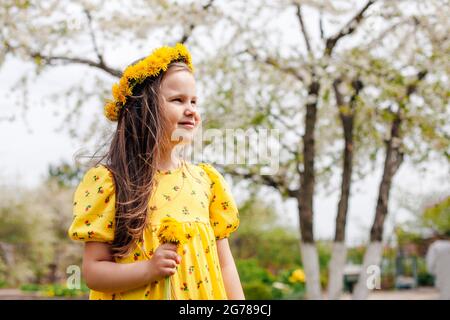 close-up portrait in profile of a smiling girl with a wreath of yellow dandelions on her head and a bouquet of dandelions in her hand with a spring Stock Photo