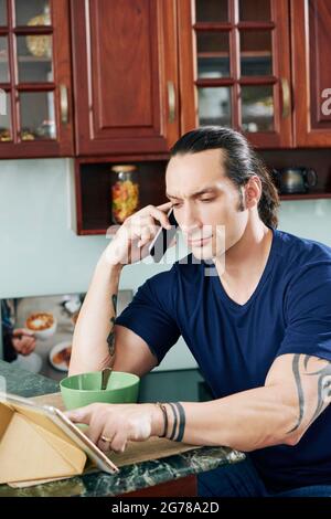 Frowning fit man sitting at kitchen counter, talking on phone with colleague and checking e-mails on tablet computer Stock Photo
