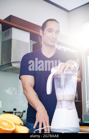 Portrait of smiling fit strong man using electronic blender when making protein cocktail with milk and bananas Stock Photo