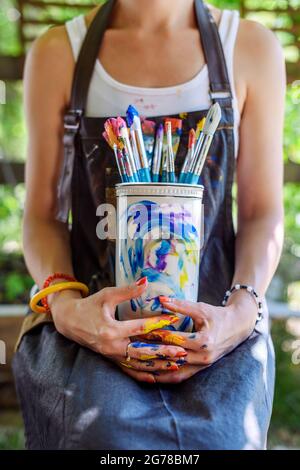 Young female painter artist holding a bunch of dirty used paintbrushes. Funky lifestyle creative hobbies background. Stock Photo