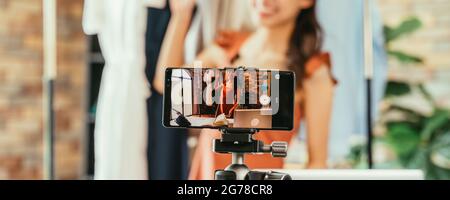 Young and beautiful Asian woman blogger showing clothes in front of smartphone camera while recording vlog video and live streaming at her shop Stock Photo