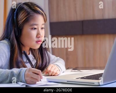 Asian girls study online with laptops. Kid wear headset typing keyboard notebook learning using internet lessons on quarantine. Students learn with in Stock Photo