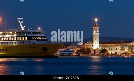Zakynthos, Zakynthos town, night shot, evening shot, harbor, front part of the Zante ferry in the middle distance, night blue water in the foreground, illuminated church of Agios Dionysios right behind the ferry, blue night sky, festive lighted church tower Stock Photo