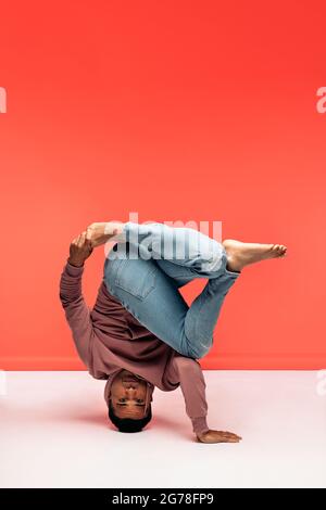 Hispanic dancer with casual clothes doing headstand and dancing in studio shot against red background. Stock Photo
