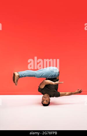 Hispanic dancer with casual clothes doing headstand and dancing in studio shot against red background. Stock Photo