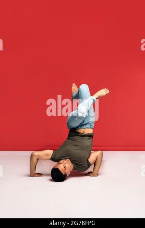 Hispanic dancer with casual clothes doing headstand and dancing in studio shot against red background. Stock Photo