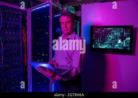 Portrait of content mature IT specialist in shirt standing with laptop at open server cabinet in data center Stock Photo