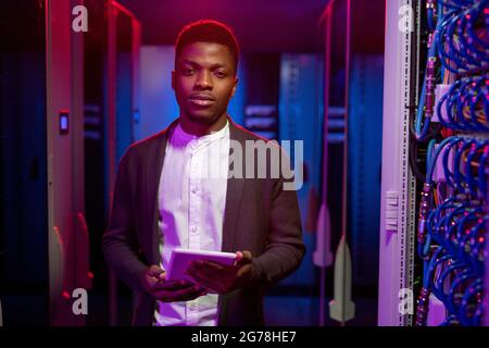 Portrait of confident handsome young African-American man responsible for managing network system standing with tablet in data center Stock Photo