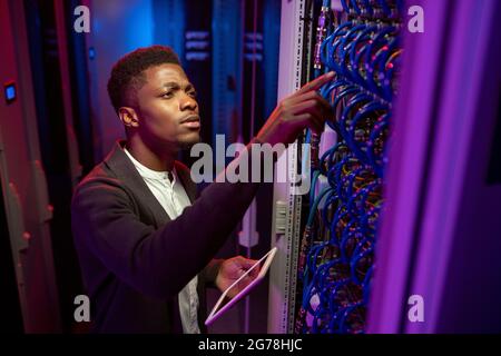 Concentrated young Black IT engineer standing with tablet in data center and checking connection of cables Stock Photo