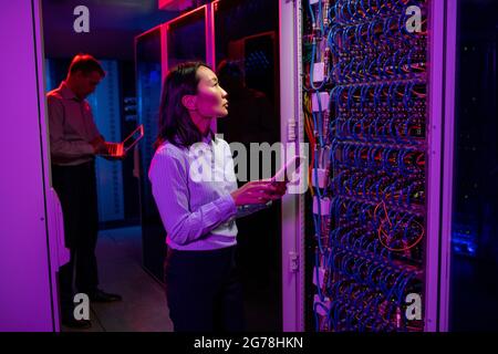 Serious young Asian IT engineer standing at server cabinet and using tablet while providing technical support at data center Stock Photo