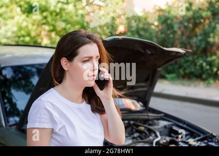 Portrait of frustrated young woman calling mobile phone for help Insurance service or ambulance after car accident. Woman in front of broken car with Stock Photo