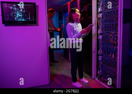 Serious young Asian network administrator standing in server room illuminated with neon light and using tablet while examining connections of cables Stock Photo