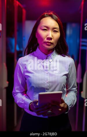Portrait of serious young Asian female server engineer in blue stripped shirt standing with tablet in data center, neon light effect Stock Photo