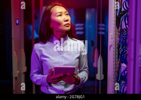 Serious pensive young Asian IT specialist in stripped shirt standing with tablet and using tablet while checking Internet connection Stock Photo