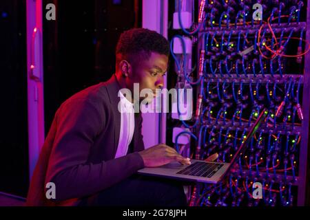 Concentrated young African-American programmer crouching near open server cabinet and using laptop while troubleshooting network system Stock Photo
