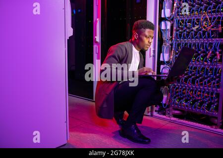 Busy young black datacenter engineer crouching in server room with neon illumination and checking network settings on laptop Stock Photo