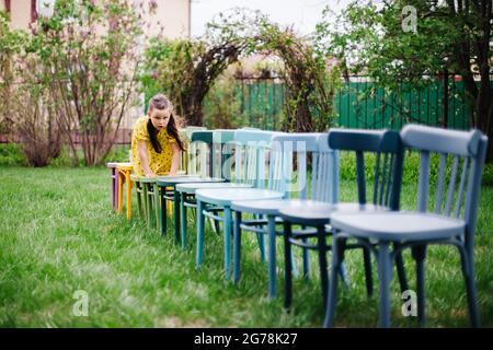 a girl crawls on all fours on a row of wooden chairs, preparing a field wedding on a green lawn in a park in nature in summer Stock Photo