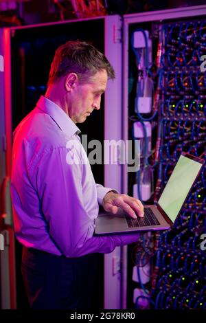 Concentrated mature Caucasian IT engineer standing against server cabinet in neon light and working with laptop Stock Photo