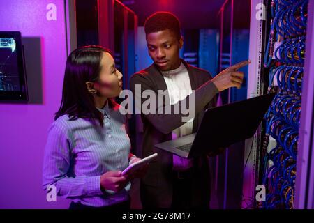 Young Afro-American technician pointing at cables while explaining server productivity to Asian colleague in data center illuminated with neon light Stock Photo