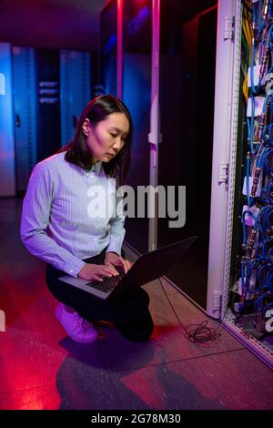 Concentrated young Asian technician crouching near server rack cabinet and using laptop while examining network connection Stock Photo