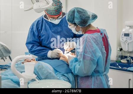 Dentist team wearing face masks and hair nets working in dental clinic with adult patient. Stock Photo