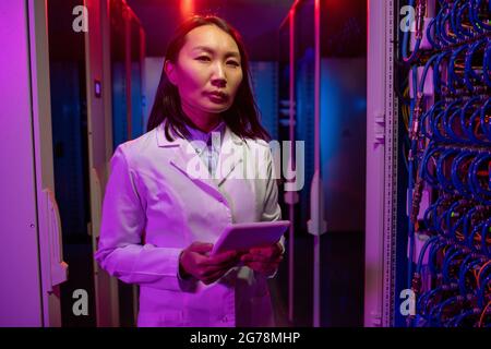 Portrait of serious young Asian support technician in white coat standing with tablet in dark neon-illuminated room Stock Photo