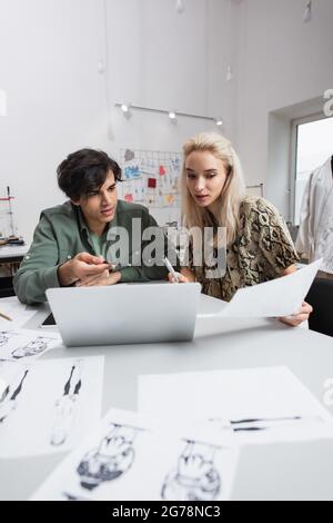 young designer pointing at laptop near blonde colleague and blurred sketches Stock Photo