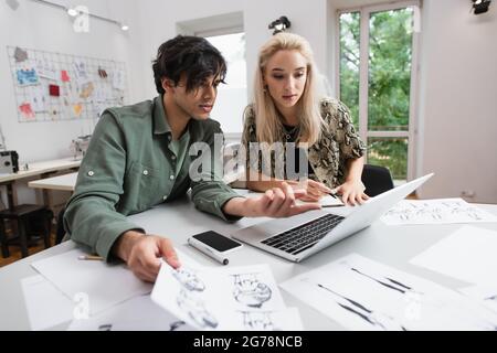 stylish designer pointing at laptop near blonde colleague and drawings at workplace Stock Photo