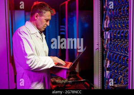 Serious concentrated mature Caucasian IT engineer standing at cart with server components and using laptop while troubleshooting server problem Stock Photo