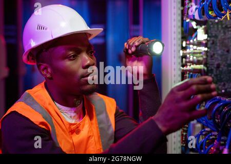 Serious young African-American server repairman in hardhat using flashlight while checking server rack Stock Photo