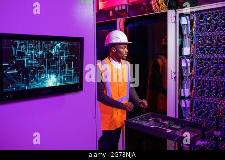Serious young Black security system engineer in hardhat and reflective vest pushing cart with server components while walking long cabinets Stock Photo