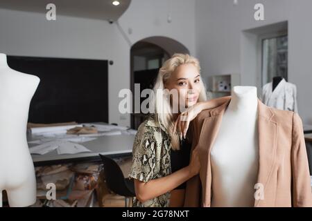 blonde fashion designer looking at camera while leaning on mannequin in atelier Stock Photo