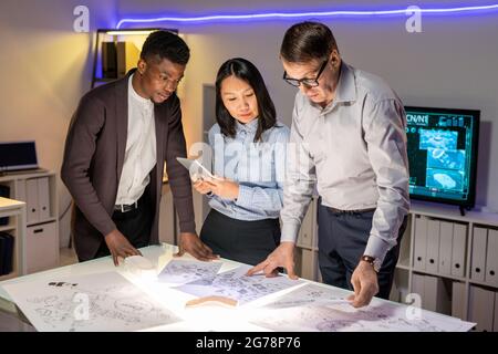 Team of concentrated interracial automotive engineers standing at table in modern office with neon illumination and discussing car sketches Stock Photo