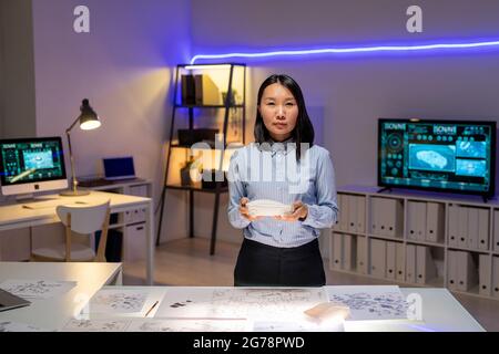 Portrait of serious young Asian female automotive exterior designer standing at desk with sketches and working with 3D model in dark office Stock Photo