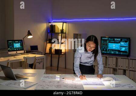 Concentrated young Asian car designer standing at table in dark office and drawing sketch while doing on deadline Stock Photo