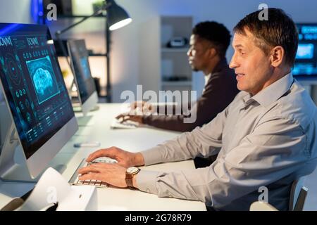 Smiling mature man sitting at table in modern office and designing car on computer against black colleague Stock Photo
