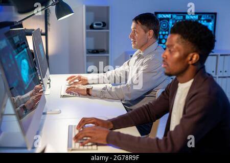 Side view of concentrated multi-ethnic industrial engineers sitting at table and typing on computer keyboards while developing design of car Stock Photo