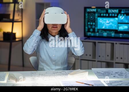 Young Asian woman in blue shirt sitting at desk with sketches and adjusting virtual reality headset while preparing for work Stock Photo