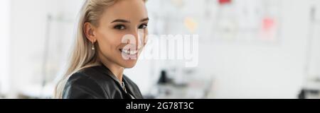 blonde fashion designer smiling at camera in tailor shop, banner Stock Photo