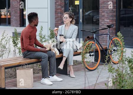 Young intercultural colleagues eating sandwiches and having coffee in urban environment Stock Photo