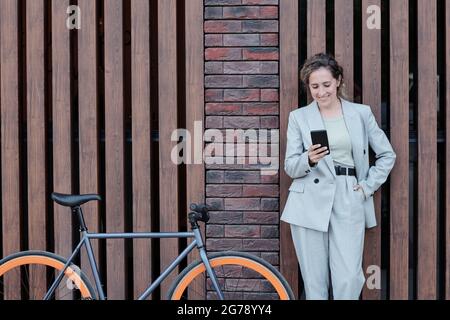 Happy elegant female scrolling in smartphone while standing by wall Stock Photo