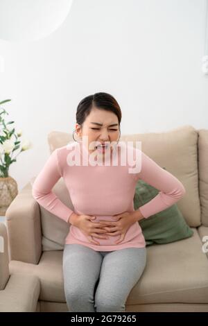 Young woman suffering from abdominal pain while sitting on sofa at home Stock Photo