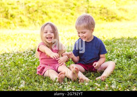 A blonde-haired boy and a girl are sitting on a green grass lawn and playing with each other. Happy childhood, fun vacation Stock Photo