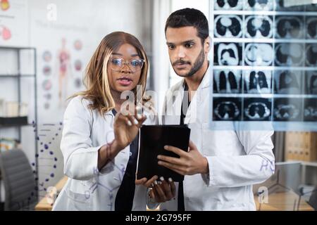 Two multiracial doctors using tablet pc while examining tomography of patient during consilium. African lady standing at modern office and writing on glass wall discussing various way of treatment. Stock Photo
