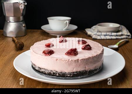 Side view of homemade delicious frozen strawberry cheesecake garnished with preserved dried strawberry in white ceramic dish with aluminum moka pot co Stock Photo