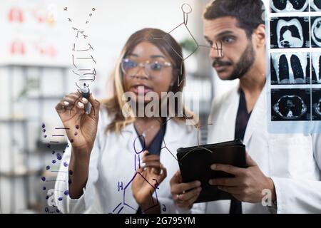 Close up portrait of two diverse scientists doctors chemists, standing together near glass wall at bright office and writing DNA structure. Focus on hand of afro woman writing on the wall Stock Photo