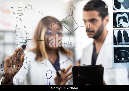 Blur view of young scientists, African woman and Arabian man, working together near glass clipboard wall, looking at the chemical structure of DNA and biological substance. Focus on hand with pen Stock Photo