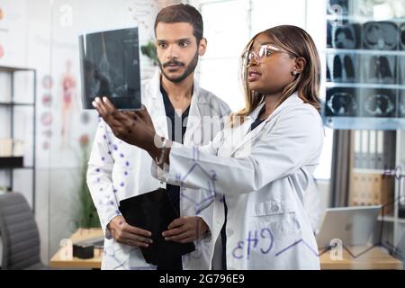 Shot of two multiracial focused young scientists standing behind glass wall in bright office room, examining patient's x-ray scan and discussing ways of treatment Stock Photo