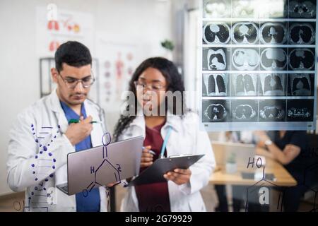 Close up view of two focused high-skilled multiethnic scientists doctors, standing near the glass wall with chemical formulas and tomography scan, using laptop. Focus on the glass wall Stock Photo